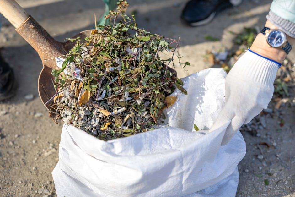 A close-up of a person wearing a white glove and a silver wristwatch, using a rusted shovel to scoop a mixture of garden debris, including small green ivy leaves, twigs, and gravel, from a white woven polypropylene sack. The sack appears to be partially filled with similar waste materials and is situated on an outdoor paved surface. The background shows some scattered leaves and dirt, with a person's foot clad in a black shoe visible in the upper left corner. The overall scene depicts a garden clearance or waste collection activity, relevant to private rubbish removal services. The lighting is natural, casting soft shadows, and the scene emphasizes the manual handling of mixed waste materials typical of on-site rubbish clearance by Waste Clearance Wandsworth, supporting alternative waste disposal methods outside local authority collections.