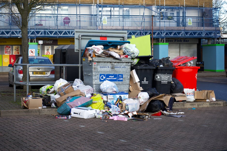A large pile of assorted rubbish and waste materials accumulated around a central metal bin in an urban parking area; the waste includes black and black plastic bags, flattened cardboard boxes, crumpled paper, and plastic packaging. The bin is labeled for mixed paper and card, and some waste appears to be overflowing, spilling onto the pavement. Behind the rubbish, there is a parked silver hatchback car with a partially obscured license plate, set against a backdrop of storefronts with yellow signage and a blue construction fence surrounding an upper-story building under renovation. The scene is illuminated by natural daylight, with a leafless tree on the left side partially framing the image. This cluttered scene illustrates the need for effective rubbish clearance services like those provided by Waste Clearance Wandsworth, highlighting the importance of timely disposal and alternative waste handling options in busy urban environments.