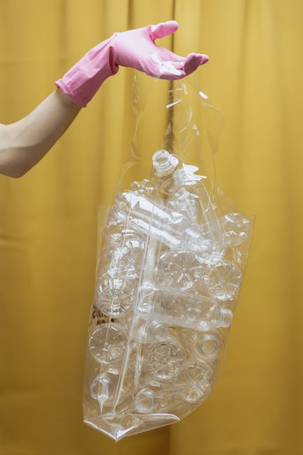 A close-up of a person wearing a white glove and a silver wristwatch, using a rusted shovel to scoop a mixture of garden debris, including small green ivy leaves, twigs, and gravel, from a white woven polypropylene sack. The sack appears to be partially filled with similar waste materials and is situated on an outdoor paved surface. The background shows some scattered leaves and dirt, with a person's foot clad in a black shoe visible in the upper left corner. The overall scene depicts a garden clearance or waste collection activity, relevant to private rubbish removal services. The lighting is natural, casting soft shadows, and the scene emphasizes the manual handling of mixed waste materials typical of on-site rubbish clearance by Waste Clearance Wandsworth, supporting alternative waste disposal methods outside local authority collections.