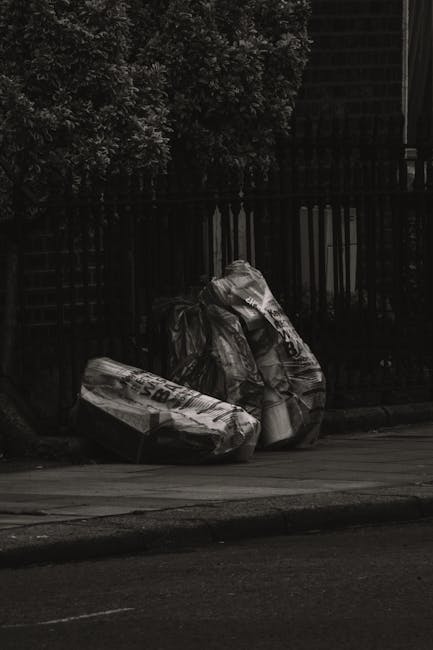 Three large bags of mixed waste, made of transparent and black plastic material, are placed on the pavement in front of a black metal fence with vertical bars. The bags appear to contain cardboard, paper, and other lightweight rubbish, with visible crumpling and folding. The scene is set outdoors in an urban environment, with a background of dense leafy greenery partially obscured by the fence. The lighting suggests a shaded area, casting subtle shadows on the street and on the rubbish bags. This image illustrates an example of private rubbish accumulation, potentially awaiting collection by a waste clearance service such as Waste Clearance Wandsworth, which offers on-site or alternative waste handling solutions away from local authority collection points.