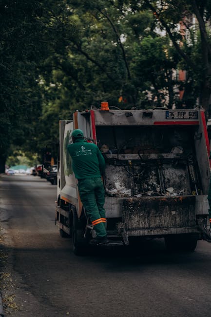 A worker dressed in green protective clothing and a helmet stands beside an open skip of a waste collection truck, positioned on a residential street with trees and parked cars in the background. The skip appears filled with burnt or charred debris, which includes blackened materials and ash. The surrounding environment includes a paved road, greenery from trees overhead, and a row of parked vehicles along the curb, suggesting a typical urban or suburban setting. The scene is captured during daylight hours, with natural light filtering through the foliage, emphasizing the industrial nature of rubbish removal and on-site waste handling, as provided by Waste Clearance Wandsworth. The image highlights the practical aspects of private waste collection services, including the loading and disposal of various refuse types, and reflects on the importance of professional rubbish clearance in maintaining community cleanliness.