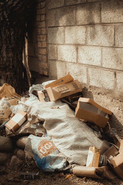 A pile of mixed waste materials resting against a weathered brick and concrete wall outdoors, consisting of flattened cardboard boxes with printed labels and logos, large white woven bags, and plastic debris. The cardboard boxes vary in size, some partially open, showing interior flaps, and are in shades of brown with printed black and orange graphics. The woven bags appear durable, textured, and slightly creased, with some partially filled, creating uneven shapes. Surrounding the waste is uneven ground with small rocks and dirt, and a dark tree trunk is visible on the left side, indicating an outdoor setting. The scene suggests a location where non-recyclable or bulky rubbish has been temporarily accumulated before collection or disposal, reflecting an environment typical for private waste clearance services like those offered by Waste Clearance Wandsworth. The lighting is natural and subdued, emphasizing the rough textures and muted tones of the waste and surrounding surface, aligning with the context of independent rubbish removal and alternative waste management options.