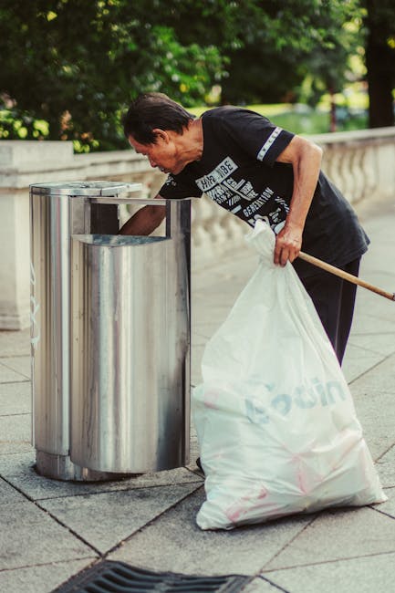 A middle-aged man with dark hair, dressed in a black T-shirt with white text and black trousers, is seen disposing of rubbish into a metallic public trash bin located on a paved outdoor area. The man leans forward, holding a white plastic bag filled with waste in his left hand, while using his right hand to lift the lid of the cylindrical, stainless steel bin, which has a shiny, reflective surface. Behind him are lush green trees and a decorative stone railing, indicating a park or street setting. The scene is well-lit with natural daylight, highlighting the textures of the plastic bag, the metal bin, and the surrounding environment. The action illustrates an individual engaging in personal waste management, consistent with private rubbish disposal or on-site clearance, supported by Waste Clearance Wandsworth's services to reduce reliance on municipal collection systems.